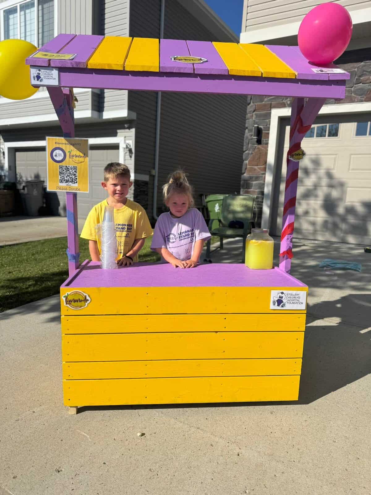 Two children standing behind a colorful lemonade stand with purple, yellow, and pink decorations during Lemonade Stand Day 2025.