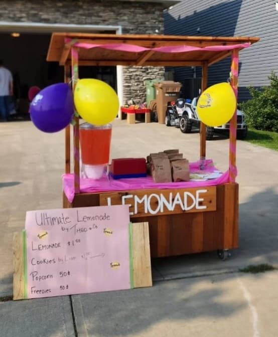 Colorful lemonade stand decorated with balloons and signs, promoting lemonade, cookies, popcorn, and freezies at Lemonade Stand Day 2025 event.