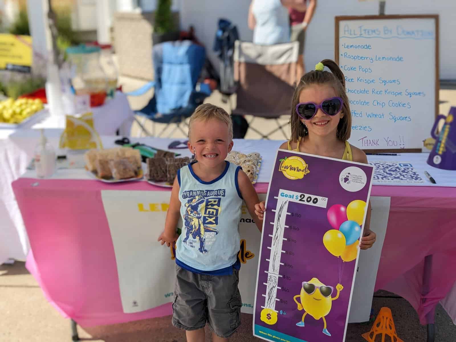 Children at Lemonade Stand Day 2025, smiling and holding a donation goal chart, promoting community fun and fundraising for a good cause.