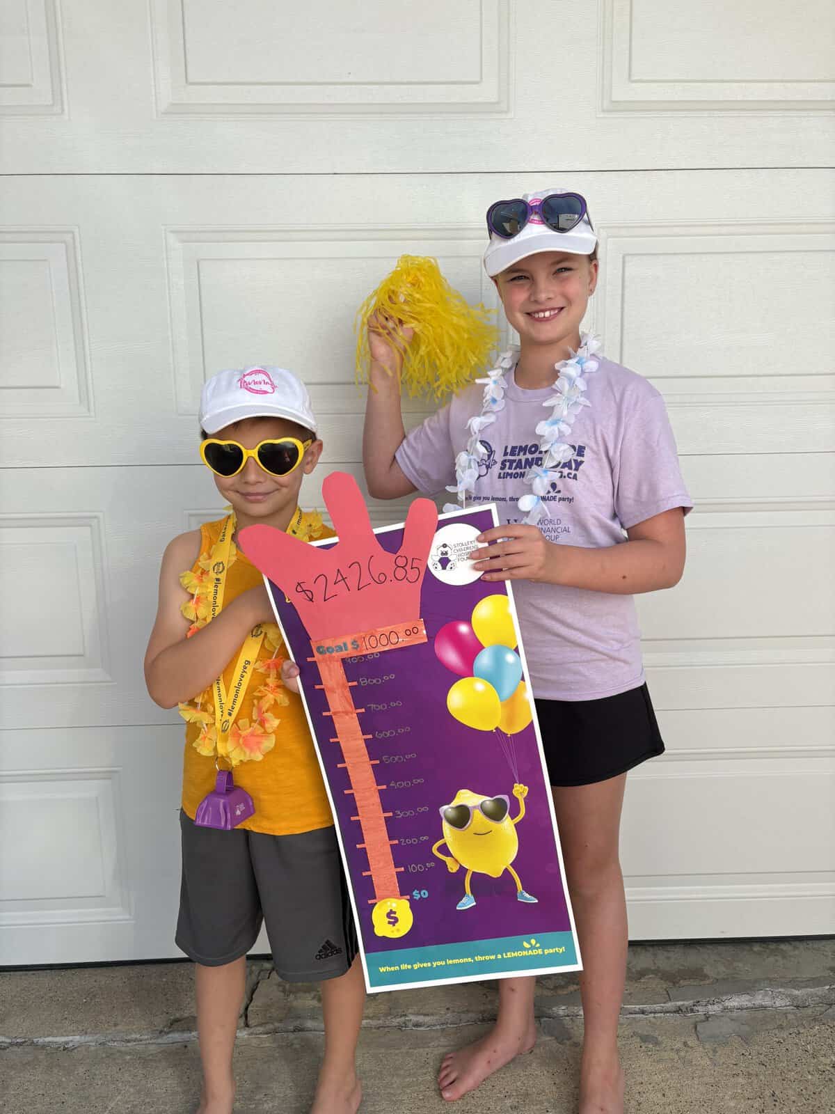 Two children at Lemonade Stand Day 2025, holding a large fundraising thermometer and a yellow pom-pom, smiling in front of a white garage door.