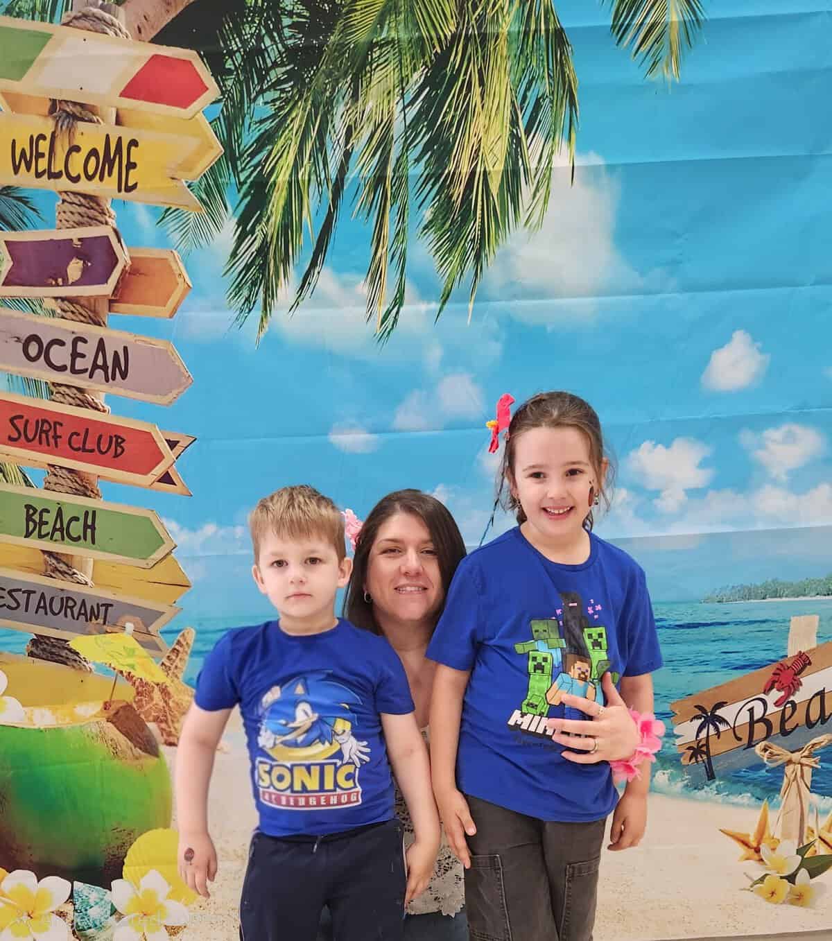 Smiling children at Lemonade Stand Day 2025 with tropical beach backdrop and colorful signs.