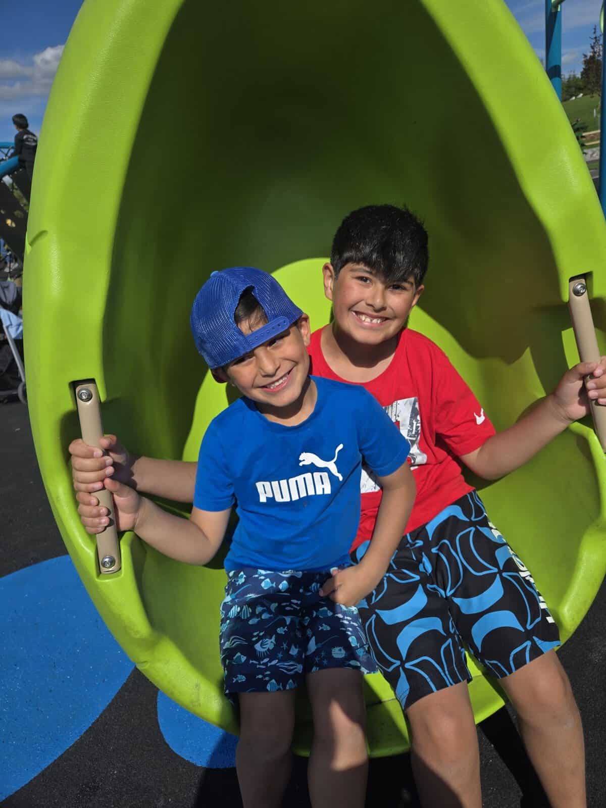 Two boys enjoying a sunny day on a green playground slide during Lemonade Stand Day 2025, smiling and having fun at the community event.