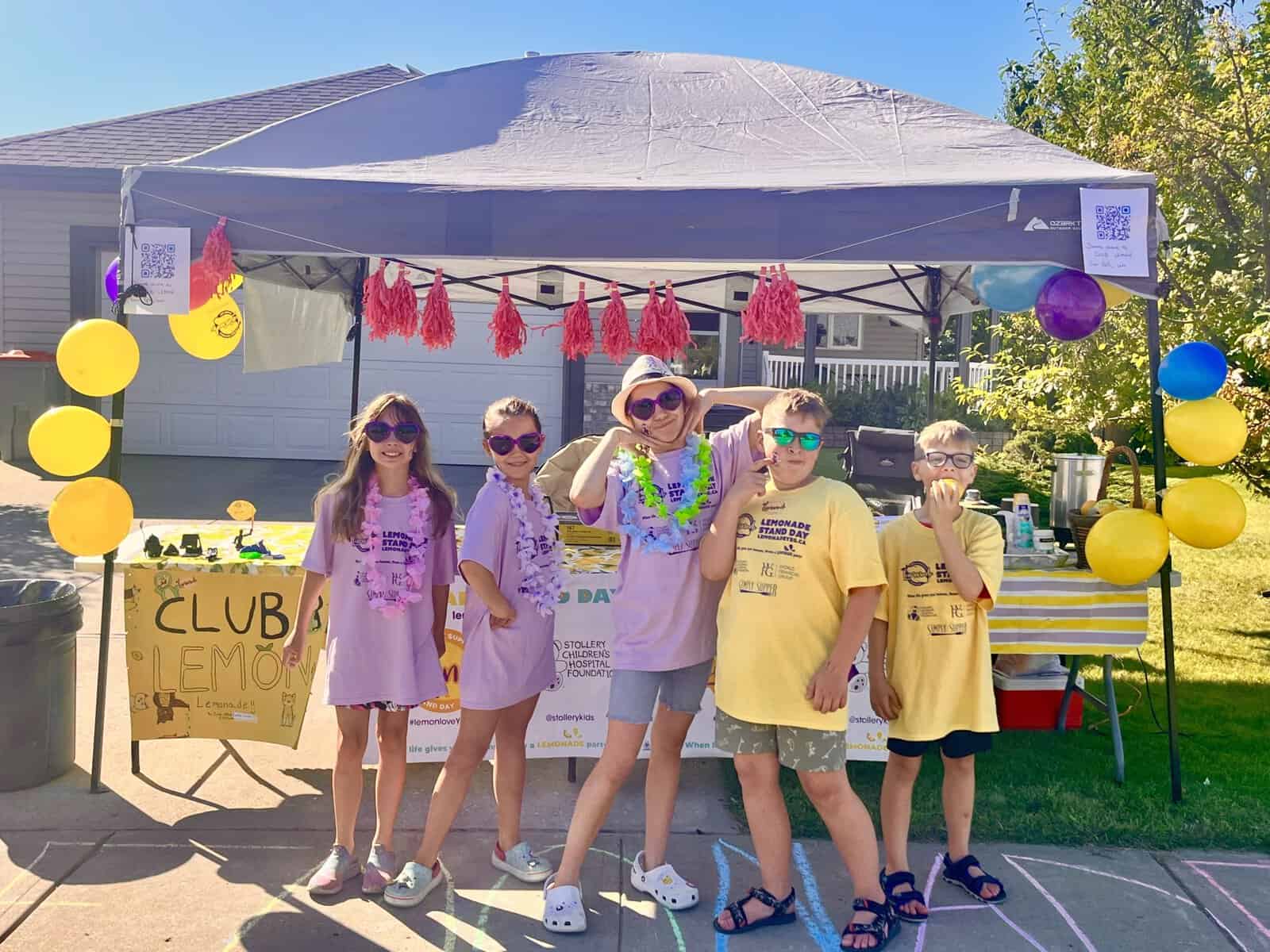 Children at Lemonade Stand Day 2025, a community event for kids to sell lemonade, with colorful decorations and a tent in a sunny outdoor setting.